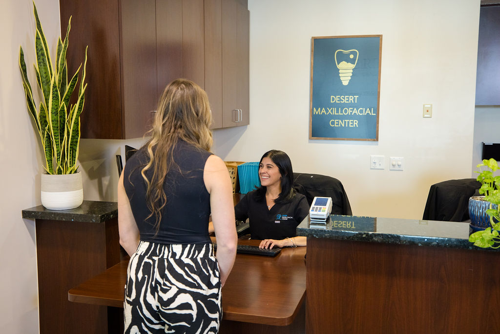 A patient at the Desert Maxillofacial Center reception desk, with the practice's sign on the wall behind it.