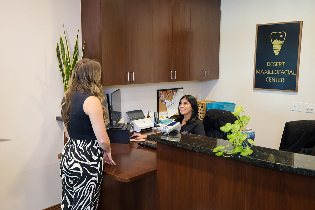A patient standing at the Desert Maxillofacial Center reception counter, speaking with a team member seated behind it.