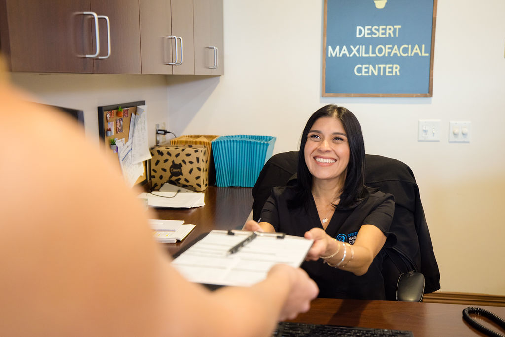 A Desert Maxillofacial Center team member at the reception desk, smiling as she hands a clipboard and pen across to a patient.