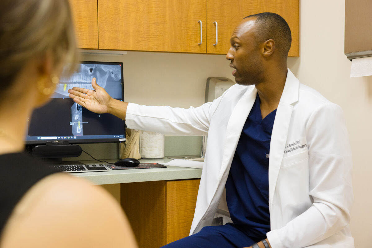 Dr. Erondu seated beside a monitor, gesturing at a dental scan while talking a patient through it.