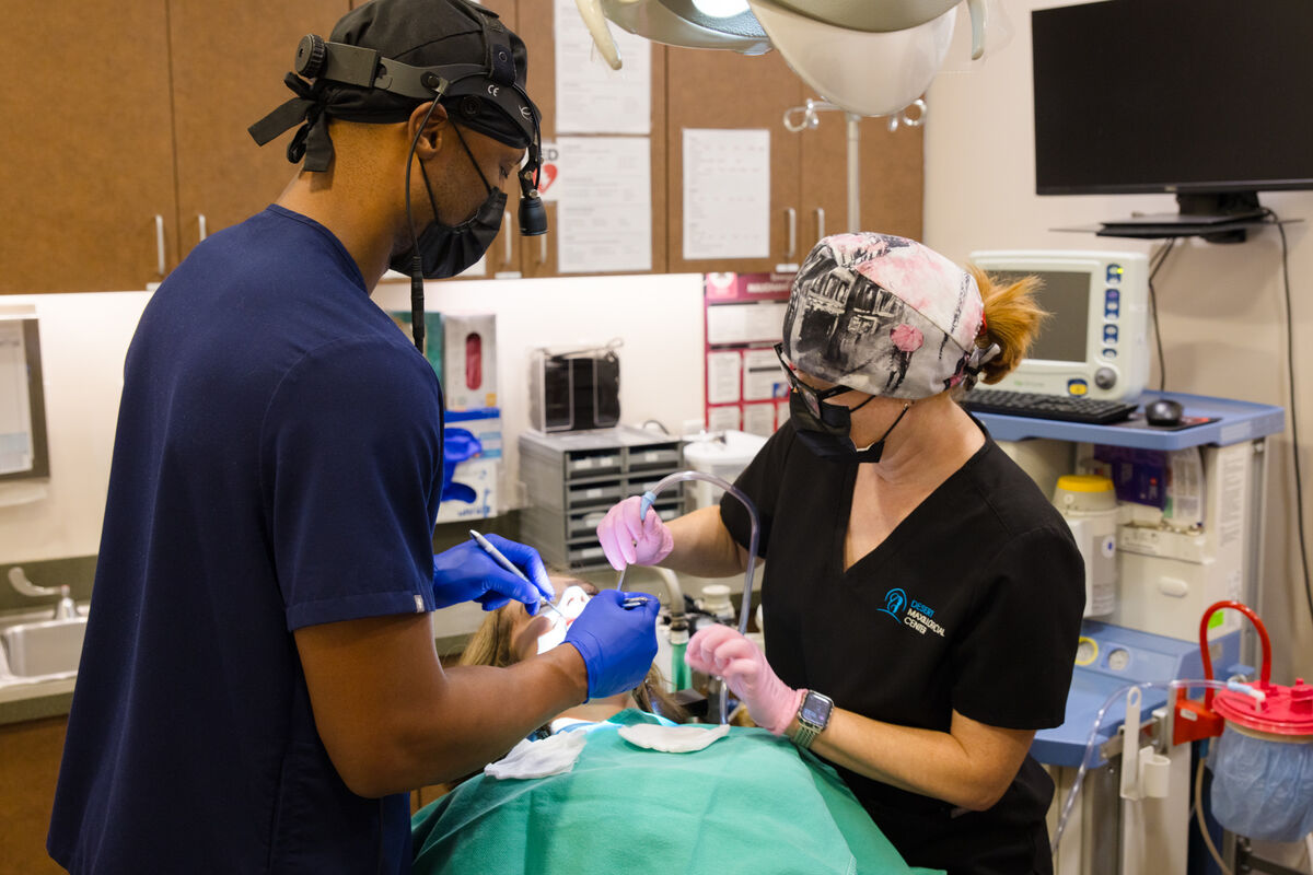 Two clinicians in surgical loupes and caps working over a draped patient in the operatory.
