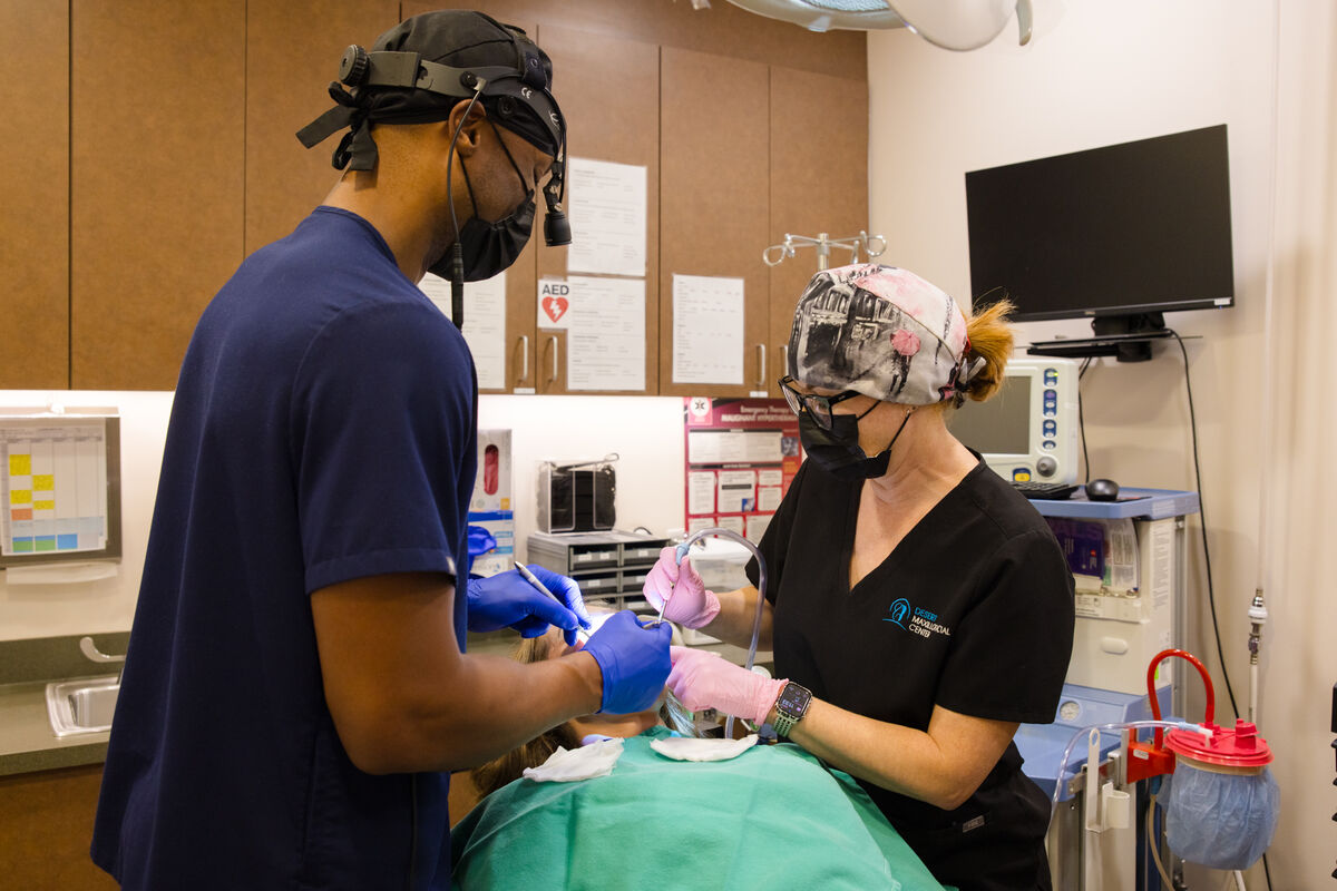 Two masked clinicians in surgical loupes working over a draped patient in the operatory.
