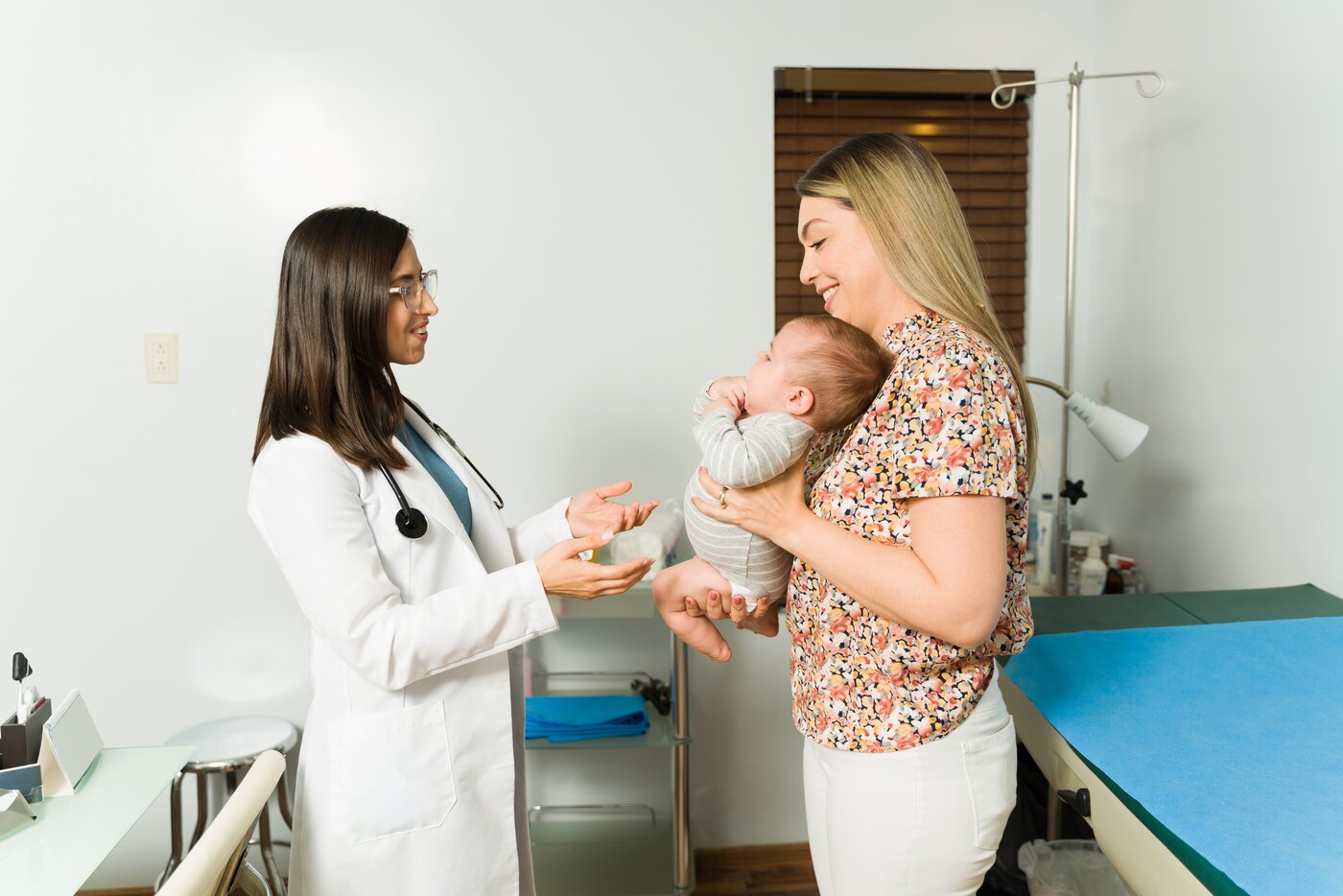 A clinician in a white coat greeting a parent who is holding an infant, in a consultation room.