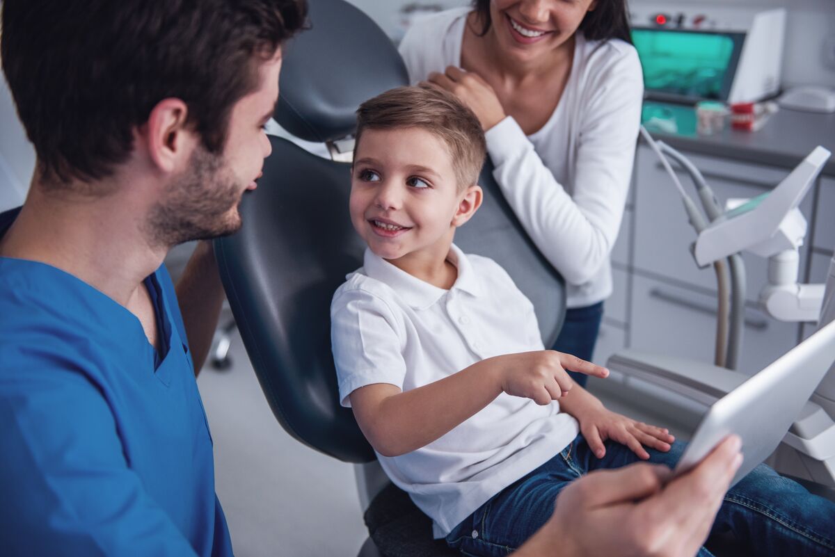 A clinician showing a tablet to a boy in the treatment chair while a parent stands beside him.