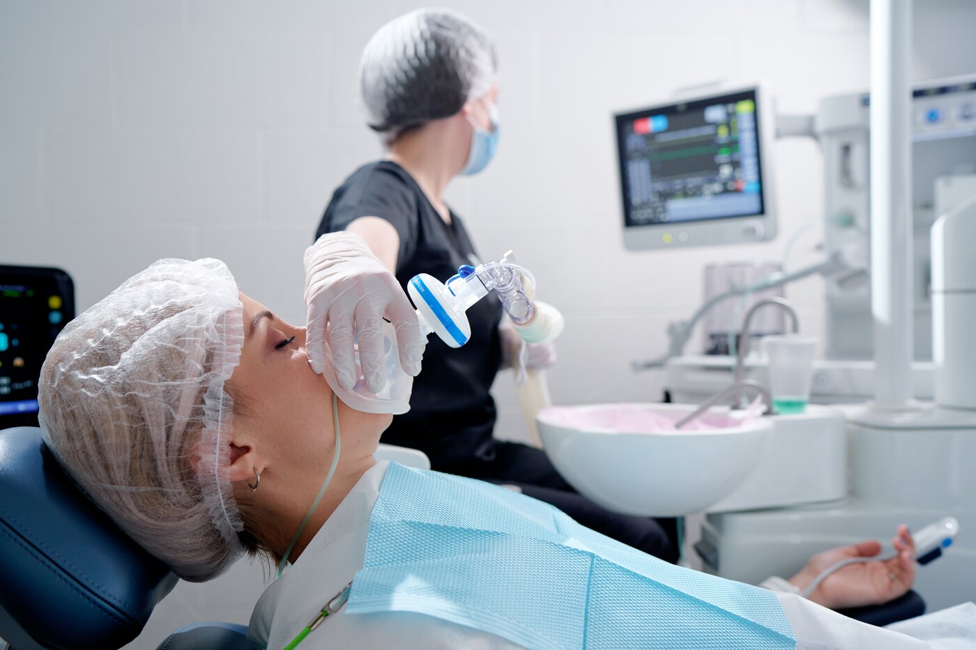 A patient reclined in a surgical chair under sedation, with a clinician and a vital-signs monitor alongside.