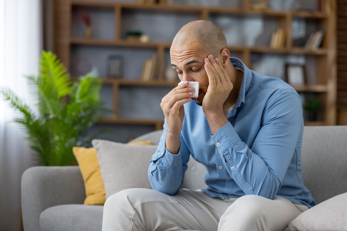 A man sitting on a sofa at home holding a tissue to his face, visibly unwell.